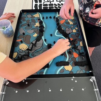 Two children placing small black figurines onto an illustrated board depicting an Aboriginal fish trap system with blue waterways and river stones.